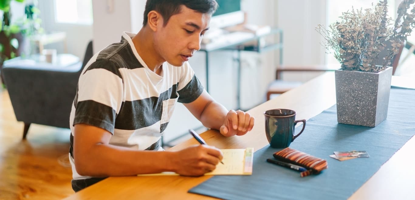 Man in striped shirt sitting at long brown table writing on yellow notepad.