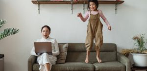 Woman working on her laptop on the sofa while her daughter jumps on the sofa