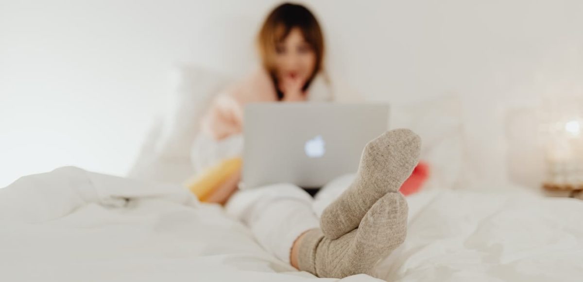 Woman in warm socks and comfy clothing sitting on bed looking at computer