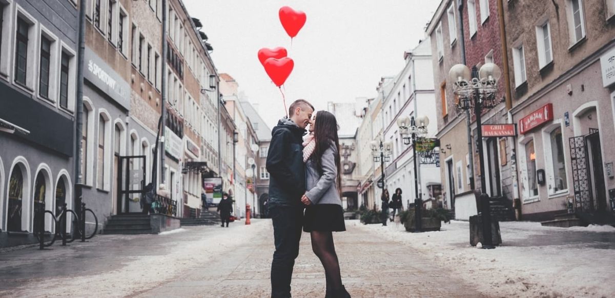 Couple embracing in middle of old town street while holding red heart baloons