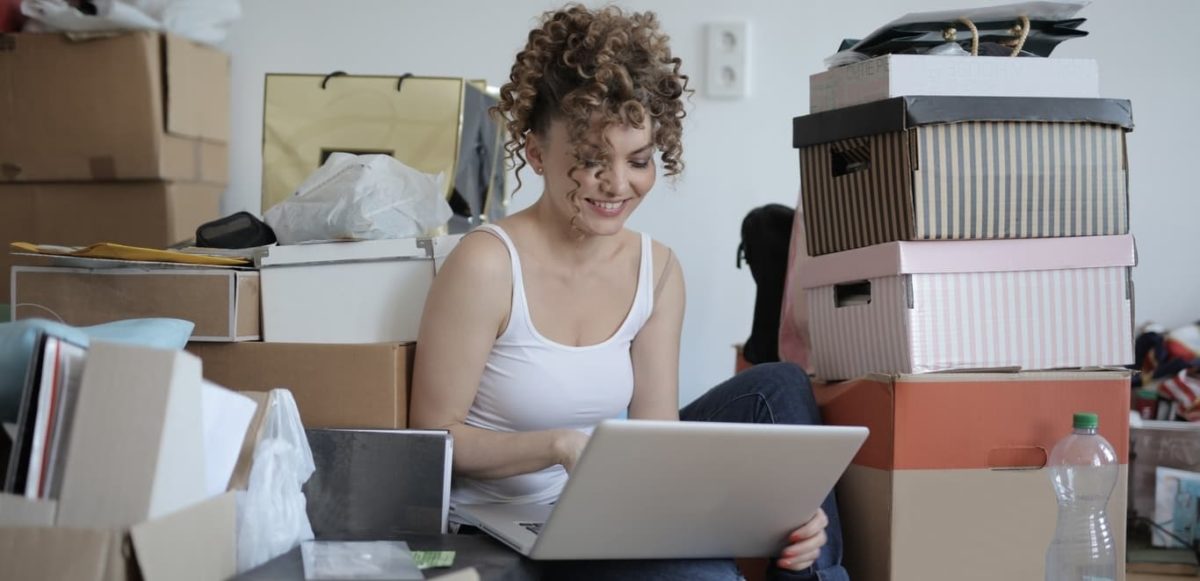 Smiling woman on laptop sitting on floor in room with many boxes and bags surrounding her.