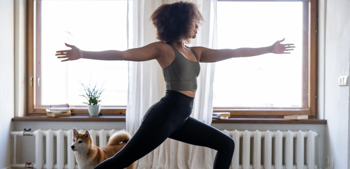 Woman doing yoga in room with wall to wall windows and a bystander dog.