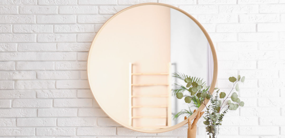 Light round wood framed decorative mirror on white brick wall above entryway table with decor and greenery.
