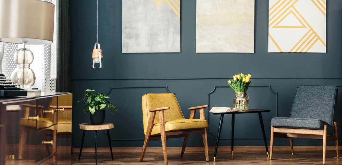 Dark accent wall with hanging artwork, a yellow chair, a blue chair, and a lamp