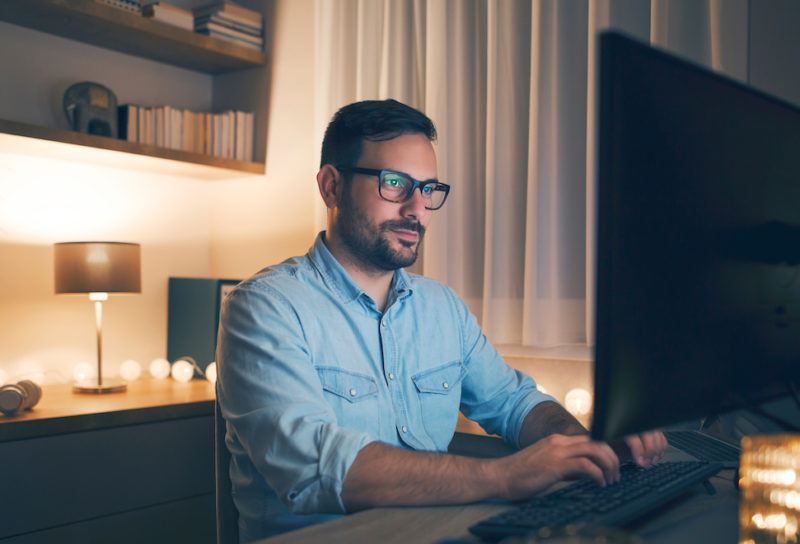 Modern young man working remotely from home at night in his basment in well decorated basement after finding basement office ideas.