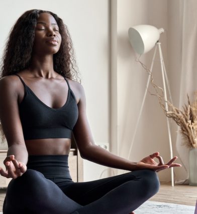 Young calm fit healthy black woman sitting on floor at home doing yoga breathing exercise, meditating learning online training virtual class on computer. Exercises for mental health concept