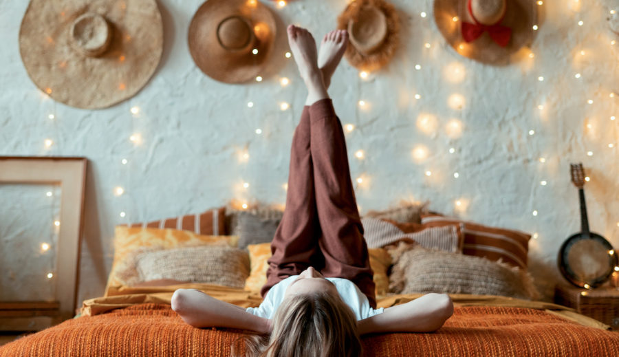 Dreamy and relaxed young adult woman lying on bed, raised legs up, resting in home at cozy bedroom in boho style