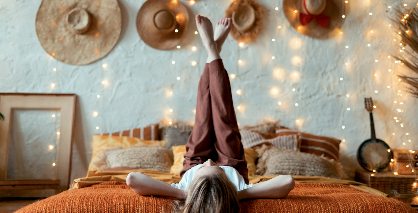 Dreamy and relaxed young adult woman lying on bed, raised legs up, resting in home at cozy bedroom in boho style