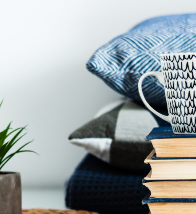 Cozy home interior decor: white and black cup, stack of books, plants in pots on a wicker stand, blue pillows on a white table in the room. Distance home education. Quarantine concept of stay home