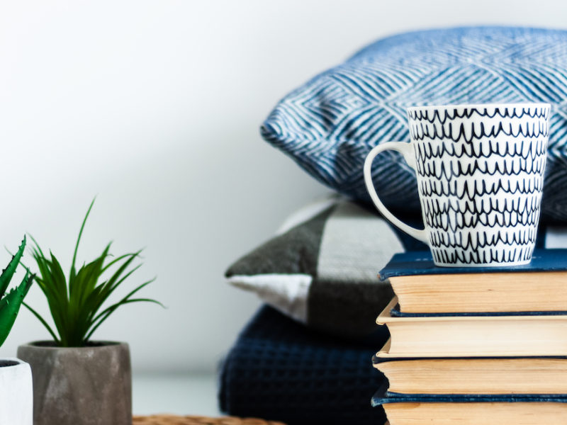 Cozy home interior decor: white and black cup, stack of books, plants in pots on a wicker stand, blue pillows on a white table in the room. Distance home education. Quarantine concept of stay home