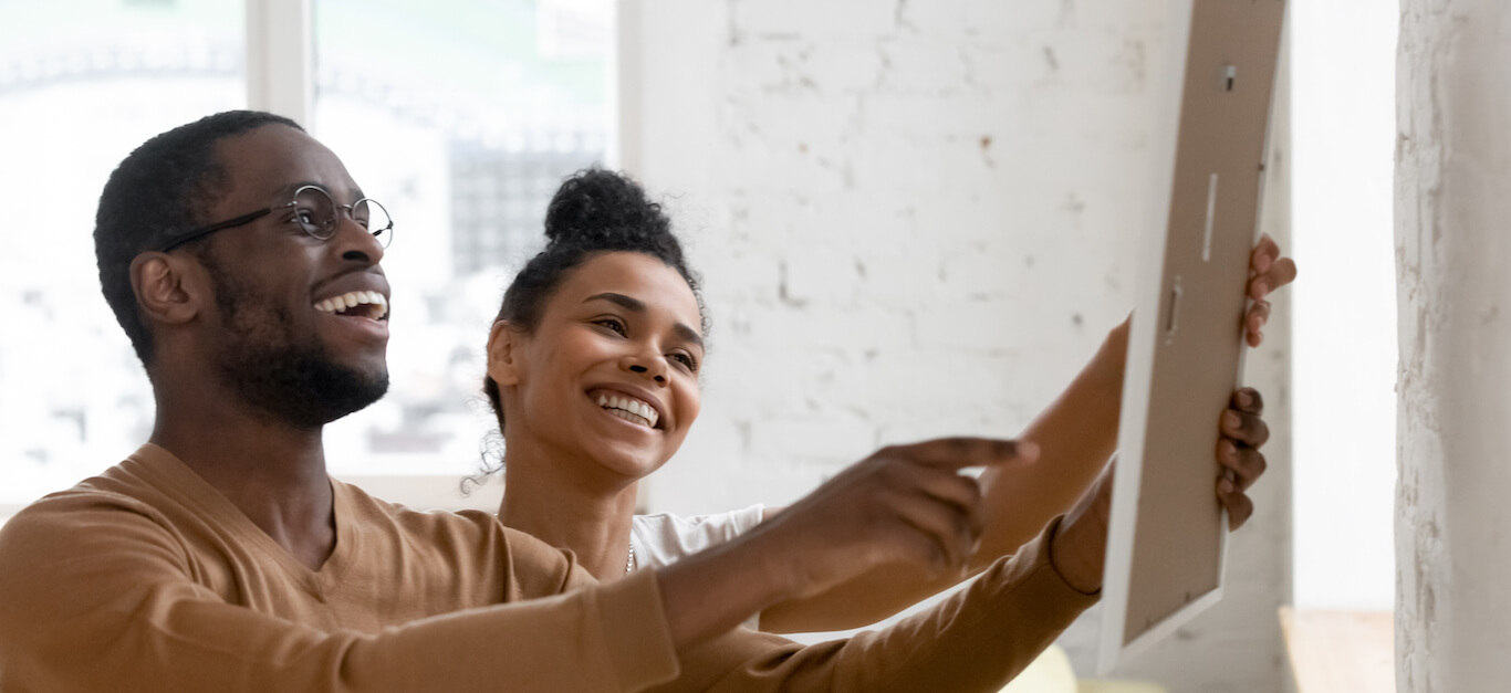 A smiling couple hangs artwork on the wall of their home.