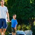 A father and son walk hand in hand, with their dog on a leash, through a neighborhood.