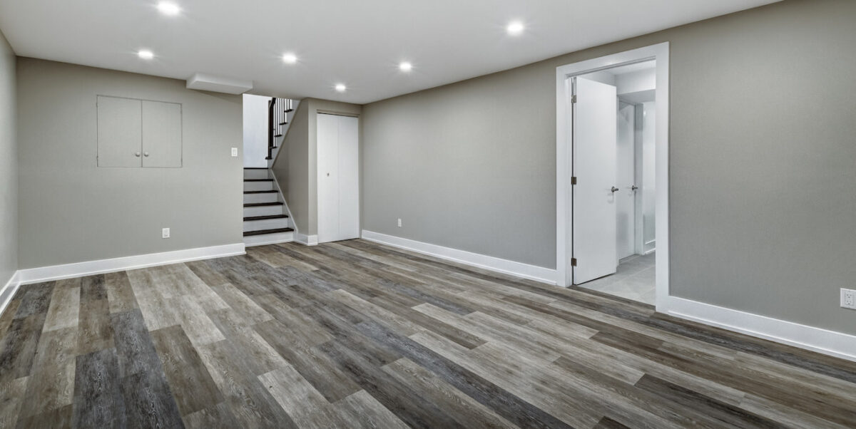 An empty finished basement with grey walls, bright overhead lighting, and grey hardwood floors.