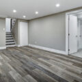 An empty finished basement with grey walls, bright overhead lighting, and grey hardwood floors.