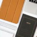 A tablet, notepad, laptop, and other home office implements evenly arranged on a white desk.