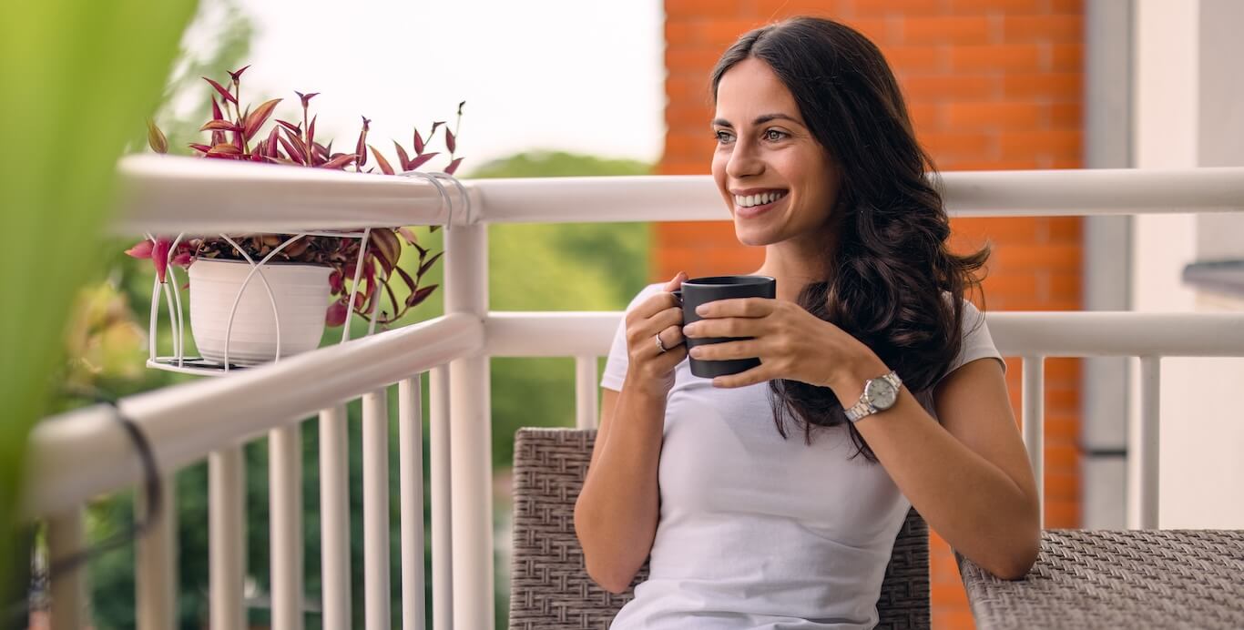 A woman enjoying a cup of coffee on her back porch, surrounded by plants and patio furniture.