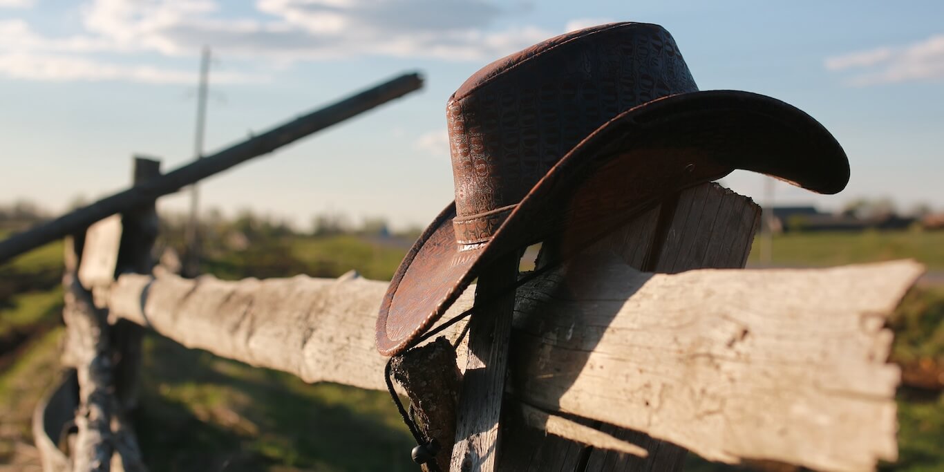 A brown cowboy hat hangs on a rustic wooden fence.