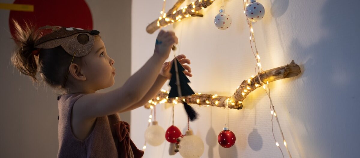 A toddler girl hanging ornaments on a tree made of lights.