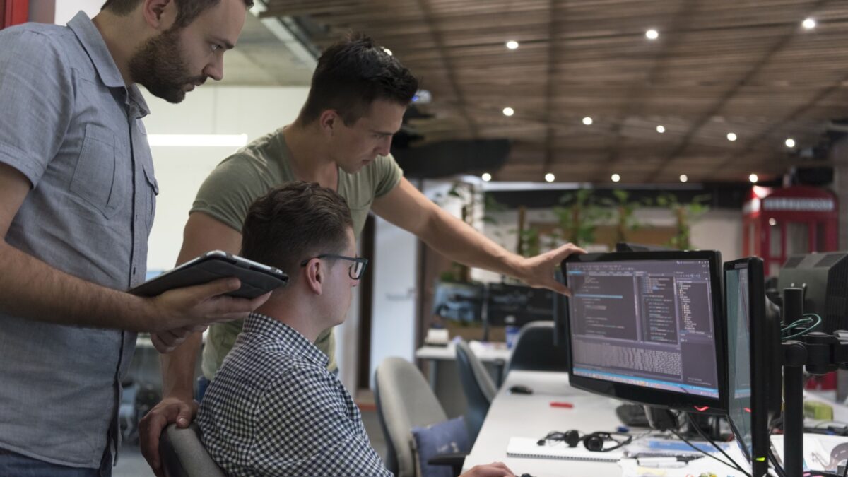 Three startup team members collaborating at a desk, reviewing code on dual monitors in a modern office setting designed with budget-friendly furnishings.