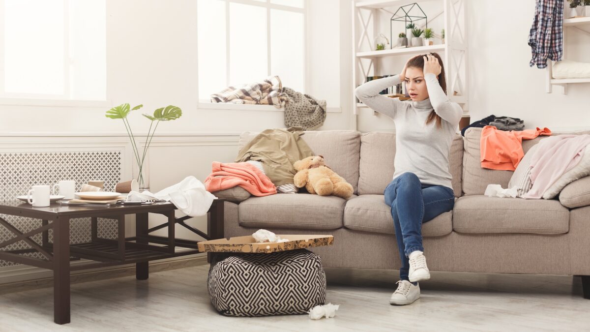 Woman on couch in cluttered room wondering how to clean a messy room and keep it clean for good. She can avoid messes and stay organized with organizational furniture at CORT Furniture Outlet.