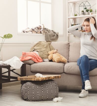 Woman on couch in cluttered room wondering how to clean a messy room and keep it clean for good. She can avoid messes and stay organized with organizational furniture at CORT Furniture Outlet.