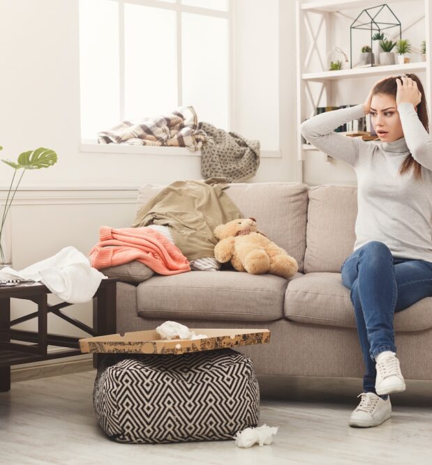 Woman on couch in cluttered room wondering how to clean a messy room and keep it clean for good. She can avoid messes and stay organized with organizational furniture at CORT Furniture Outlet.