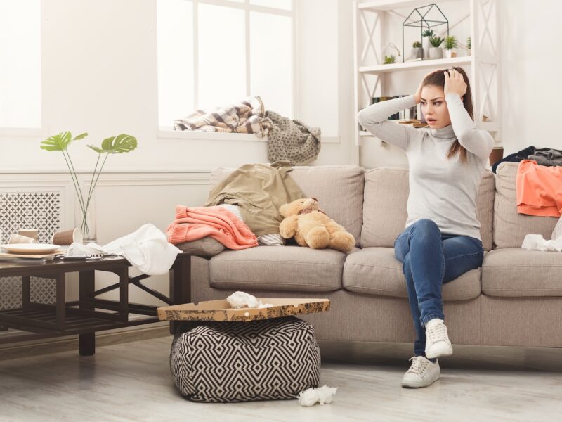 Woman on couch in cluttered room wondering how to clean a messy room and keep it clean for good. She can avoid messes and stay organized with organizational furniture at CORT Furniture Outlet.