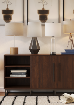 Side table in stylish neutral living room with different accent light table lamps from CORT Furniture Outlet across the surface, a stylish patterned rug, faux plants, and modern wall art ideas depicting light bulbs.